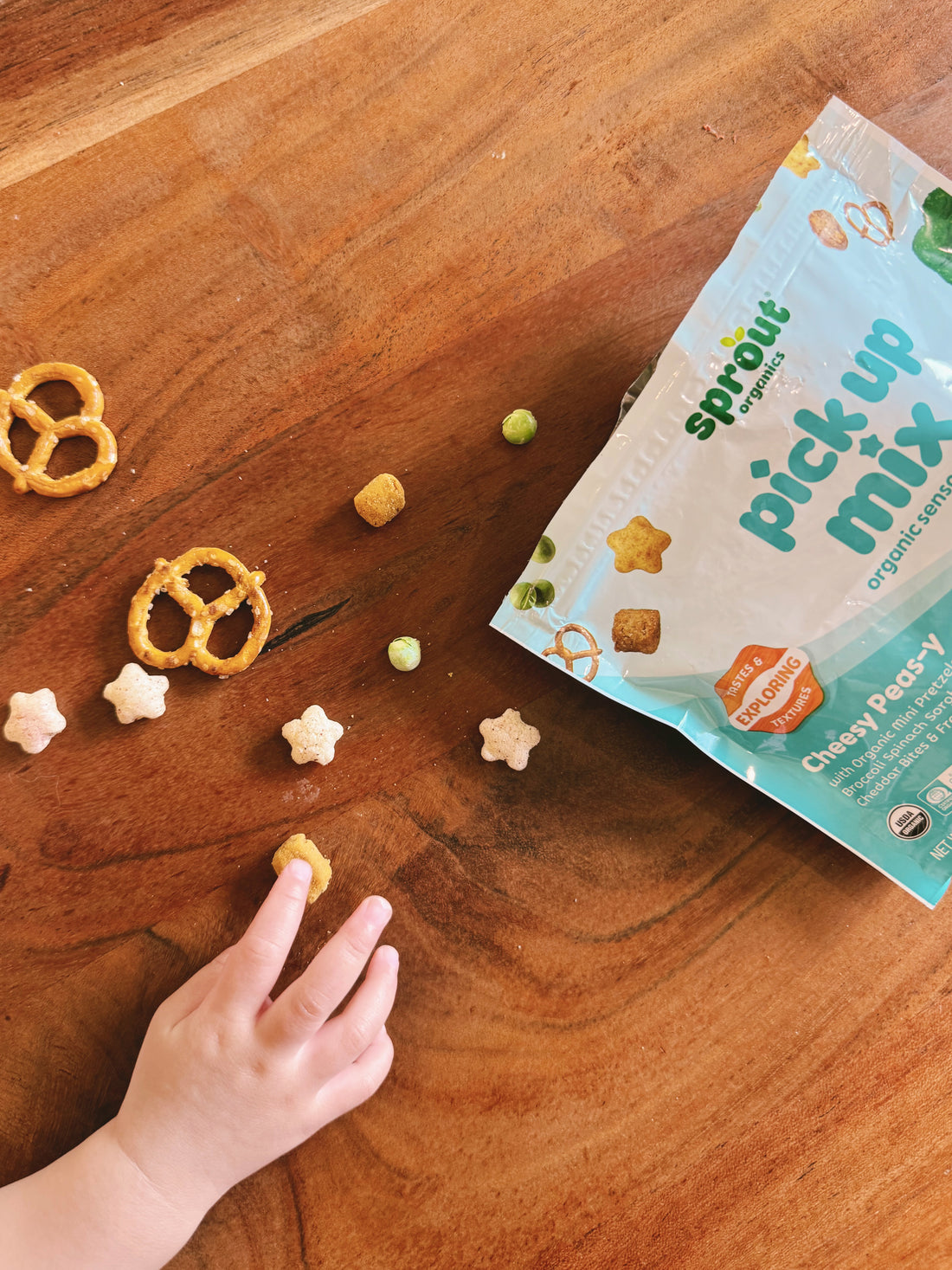 A child's hand reaches for a piece of cheddar bite snack mix from a green and white bag labeled 'sprout organics pick up mix' on a wooden tabletop, with pretzels, star-shaped puffs, and pea pieces scattered nearby.