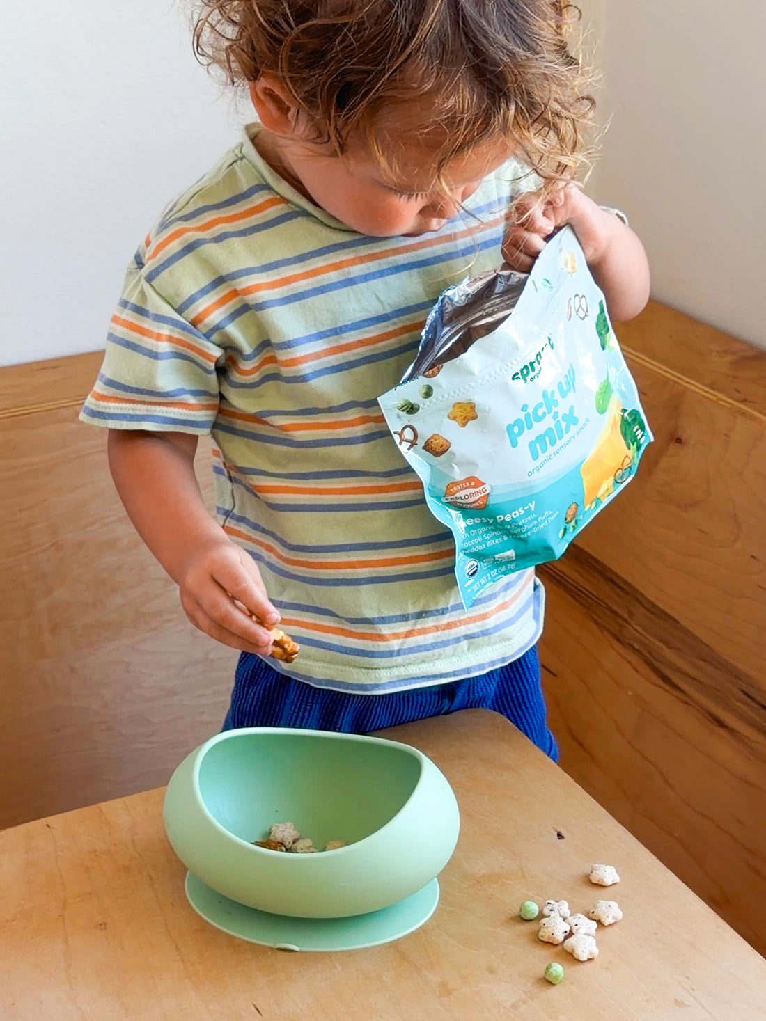 A young child wearing a striped t-shirt and blue shorts stands at a wooden dining table, holding an open bag of 'pick up mix' next to a mint green silicone suction bowl.