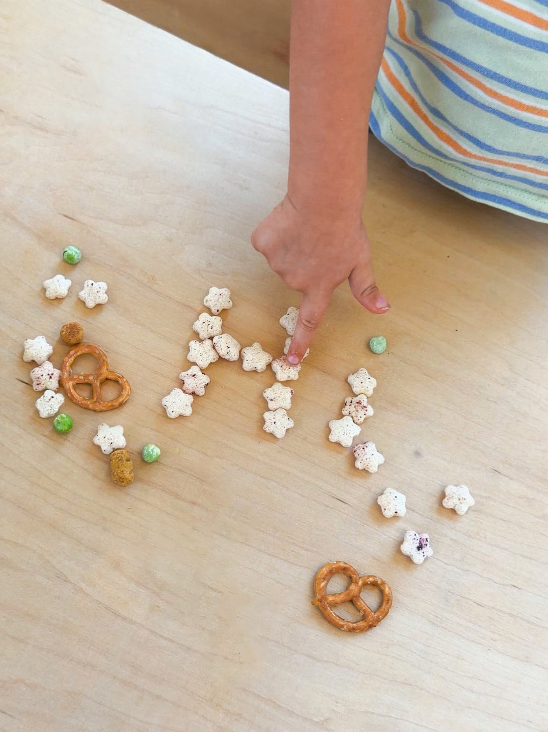 A child's hand arranging small snack items, including pretzels and veggie puffs shaped like stars, on a light wood table. The child is wearing a striped shirt with blue, orange, and white stripes.