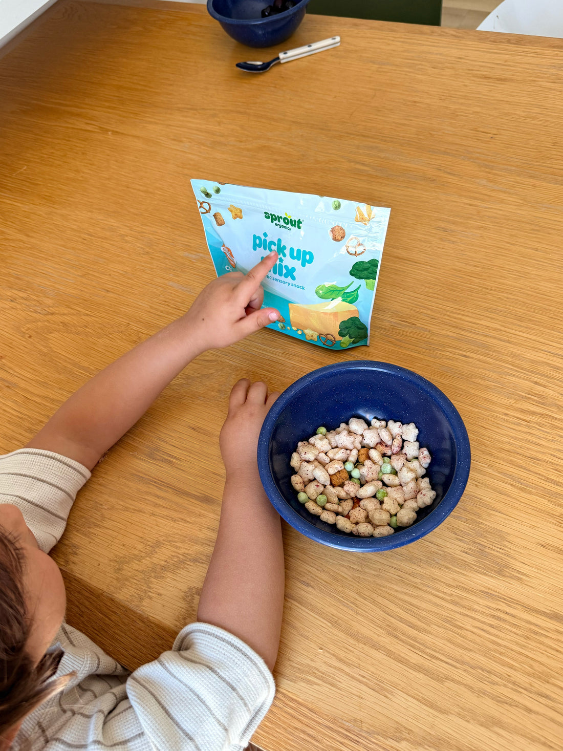 A child's hand holds a package of Sprout Pick Up Mix next to a blue bowl filled with the snack mix on a wooden table.